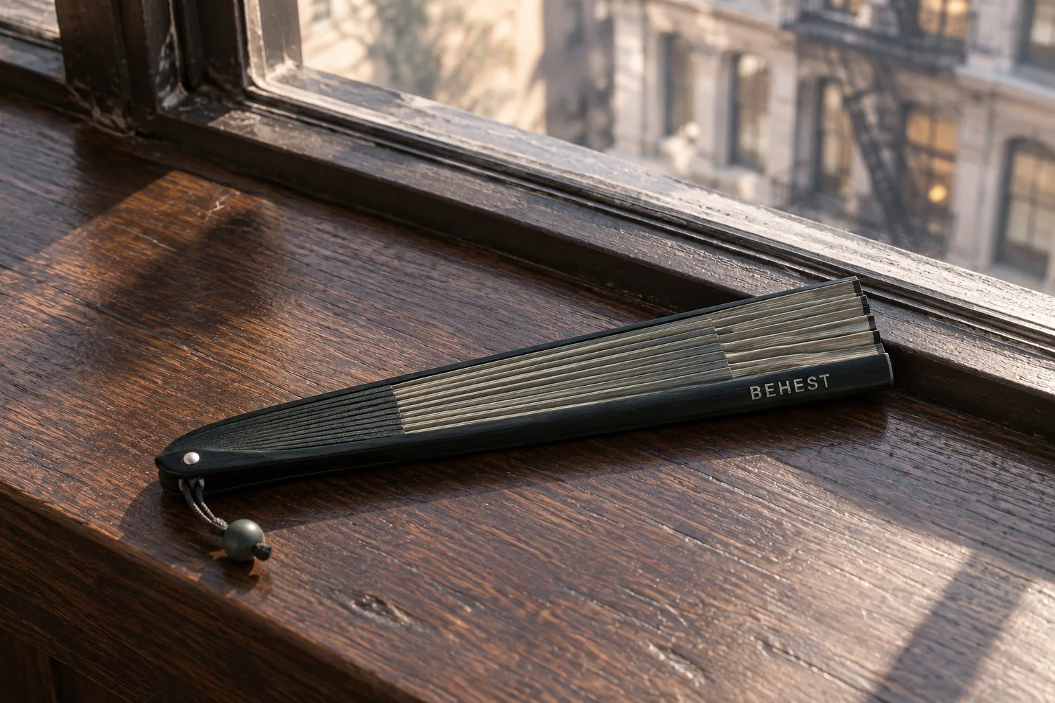 Closed folding fan with BEHEST on the guard, resting on a wooden window sill above a city street. Design concept.