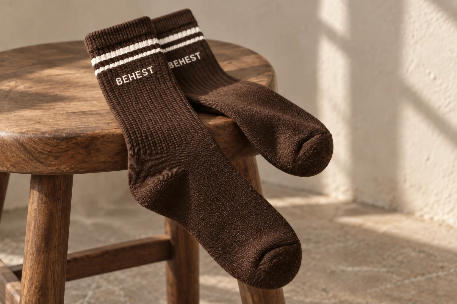 Ribbed brown BEHEST socks with two cream stripes draped over a walnut stool. Design concept.